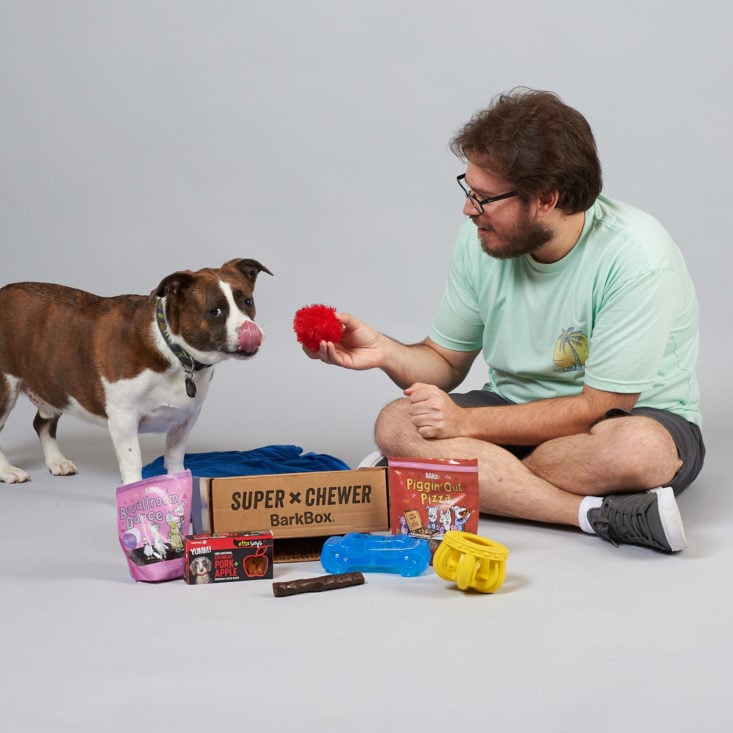 Eric unboxing the Super Chewer Box, offering a toy to a dog who's licking his nose.
